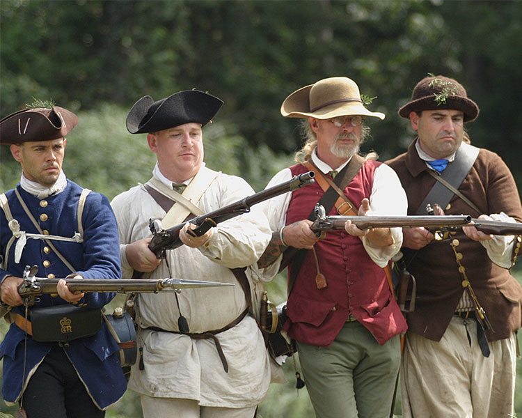 Actors dressed as colonial militia march in formation with their rifles ready to fire.