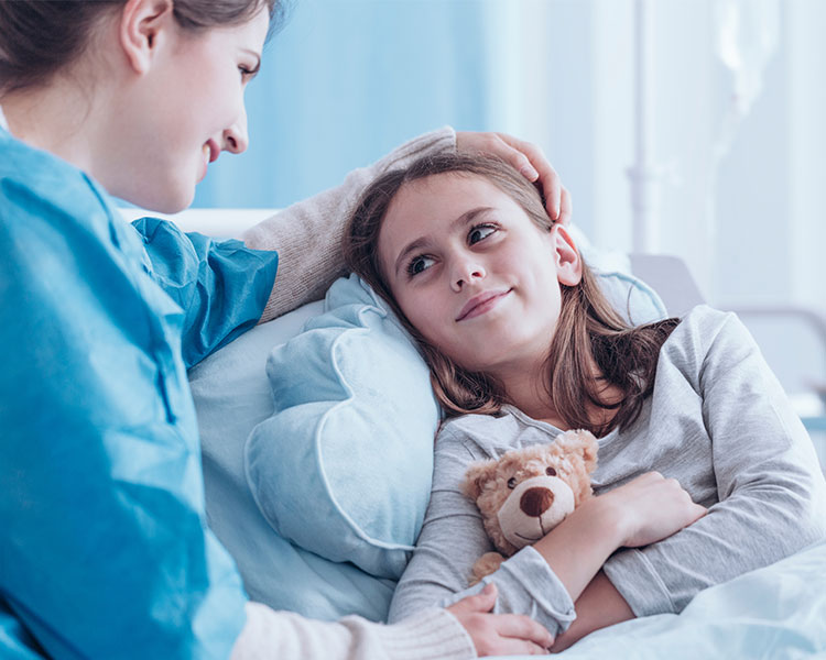 A young girl in bed smiles at her mother while holding a teddy bear