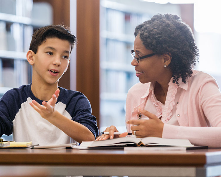 A student and tutor sitting at a desk with open books while having a conversation