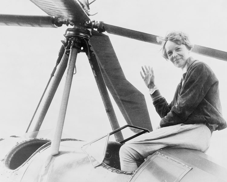 Black & white photo of Amelia Earhart sitting on her plane and waving